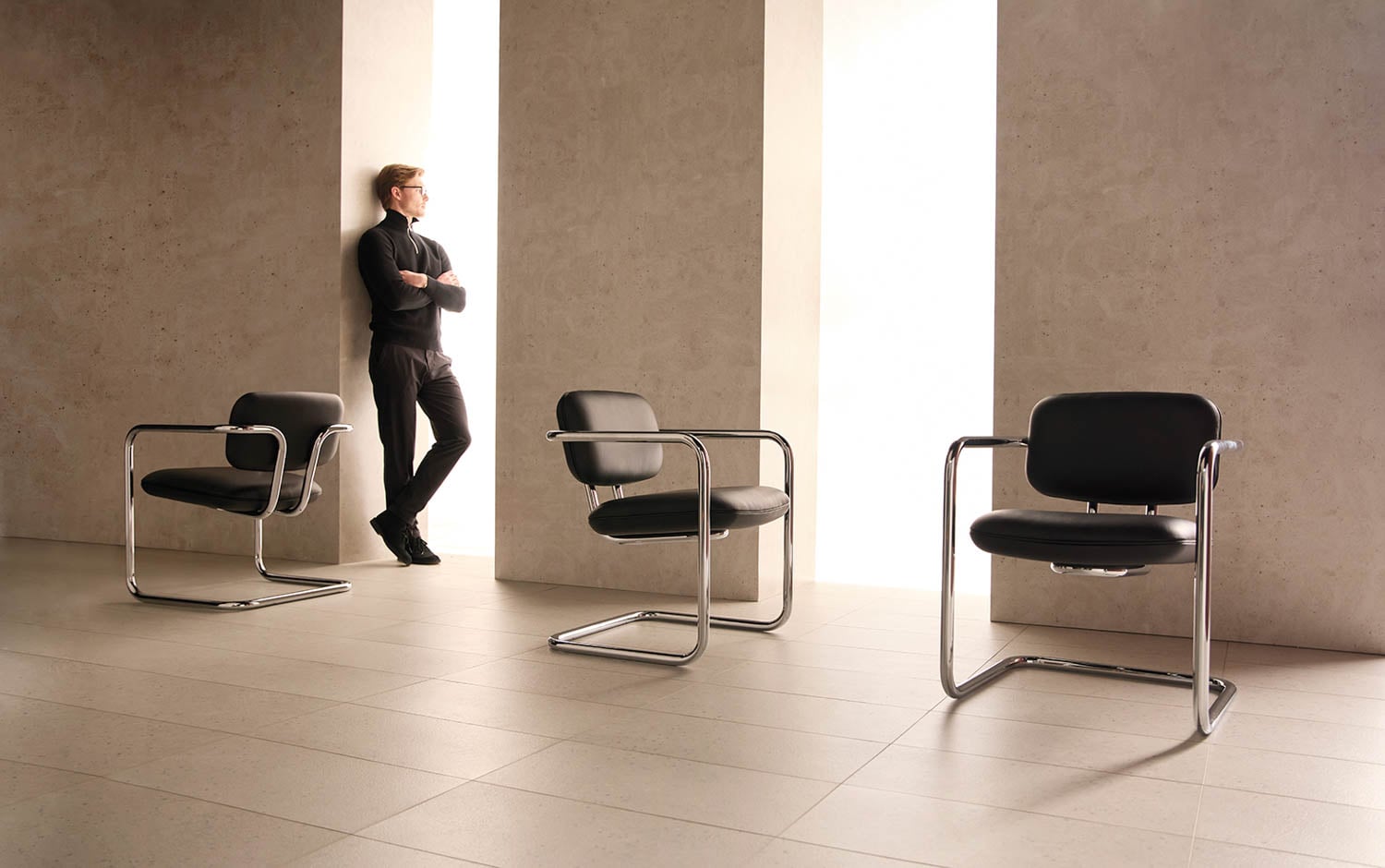 A person stands by a tall window in a modern room with beige walls and floor, next to two black chairs with metal frames.