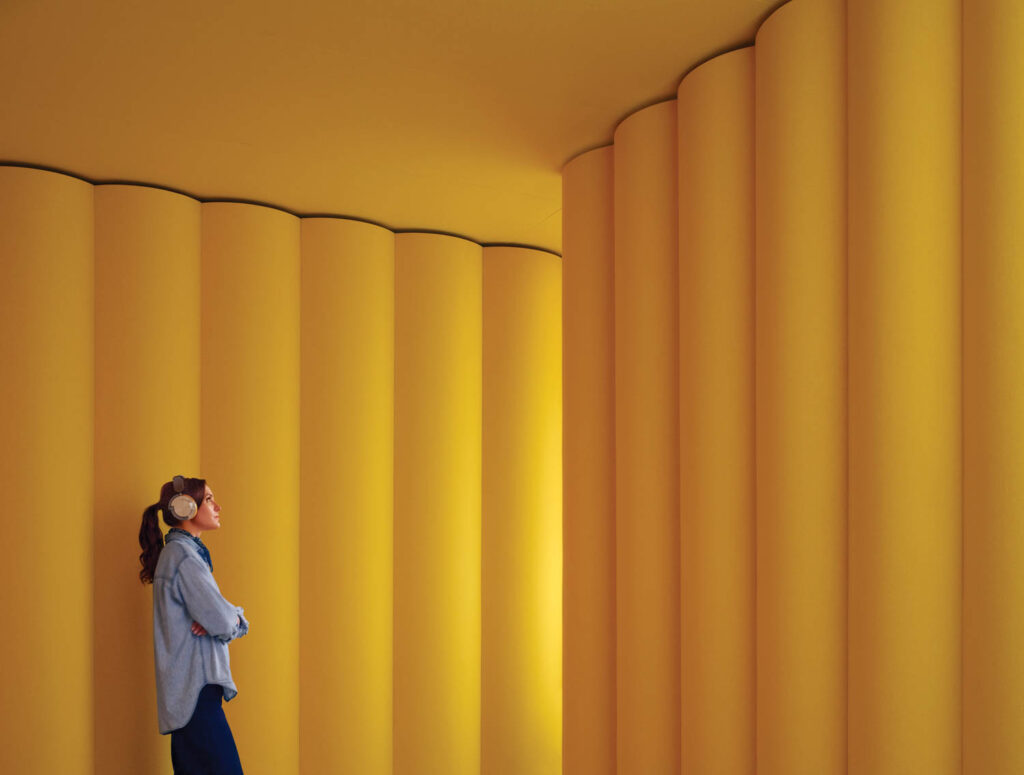 A woman wearing headphones stands against a curved yellow wall with vertical cylindrical panels, looking upward.