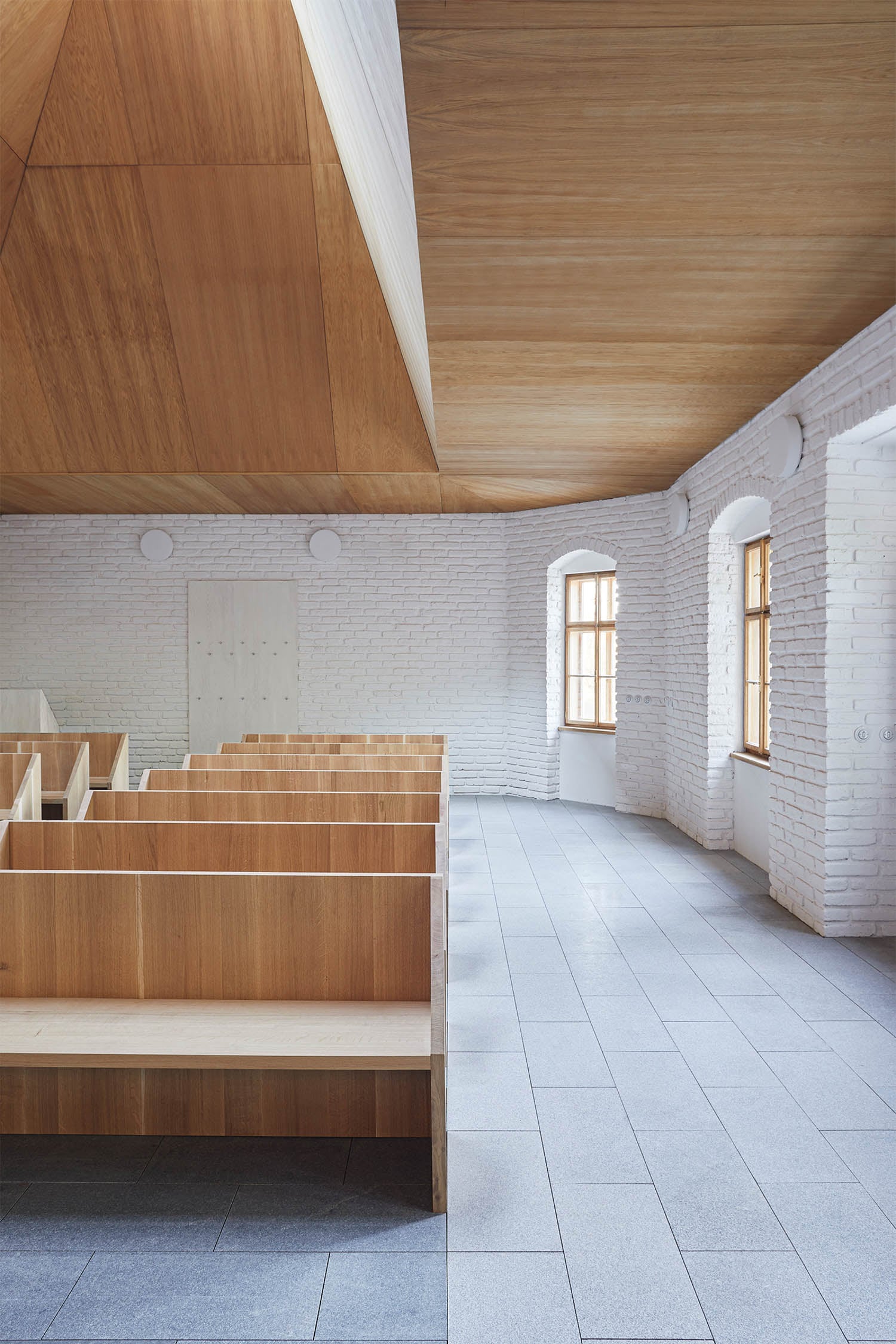 A modern interior with wooden pews, white brick walls, large windows, and a geometric wooden ceiling.
