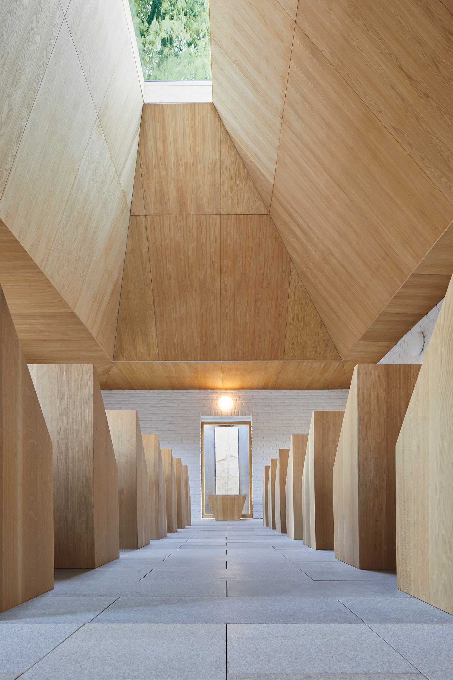 Interior view of a modern minimalist chapel with wooden pews, angled wooden ceiling panels, and a skylight above, facing a door framed by a white wall.
