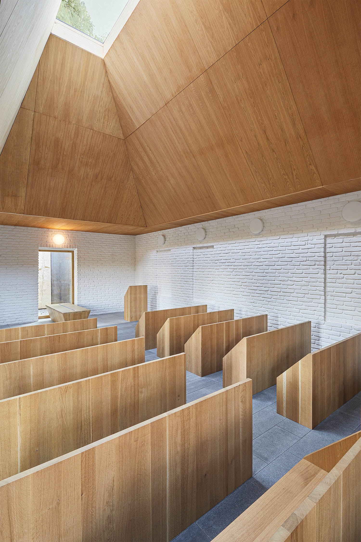 Interior of a modern chapel with wooden pews, white brick walls, grey floor tiles, and a high wooden ceiling with skylight.