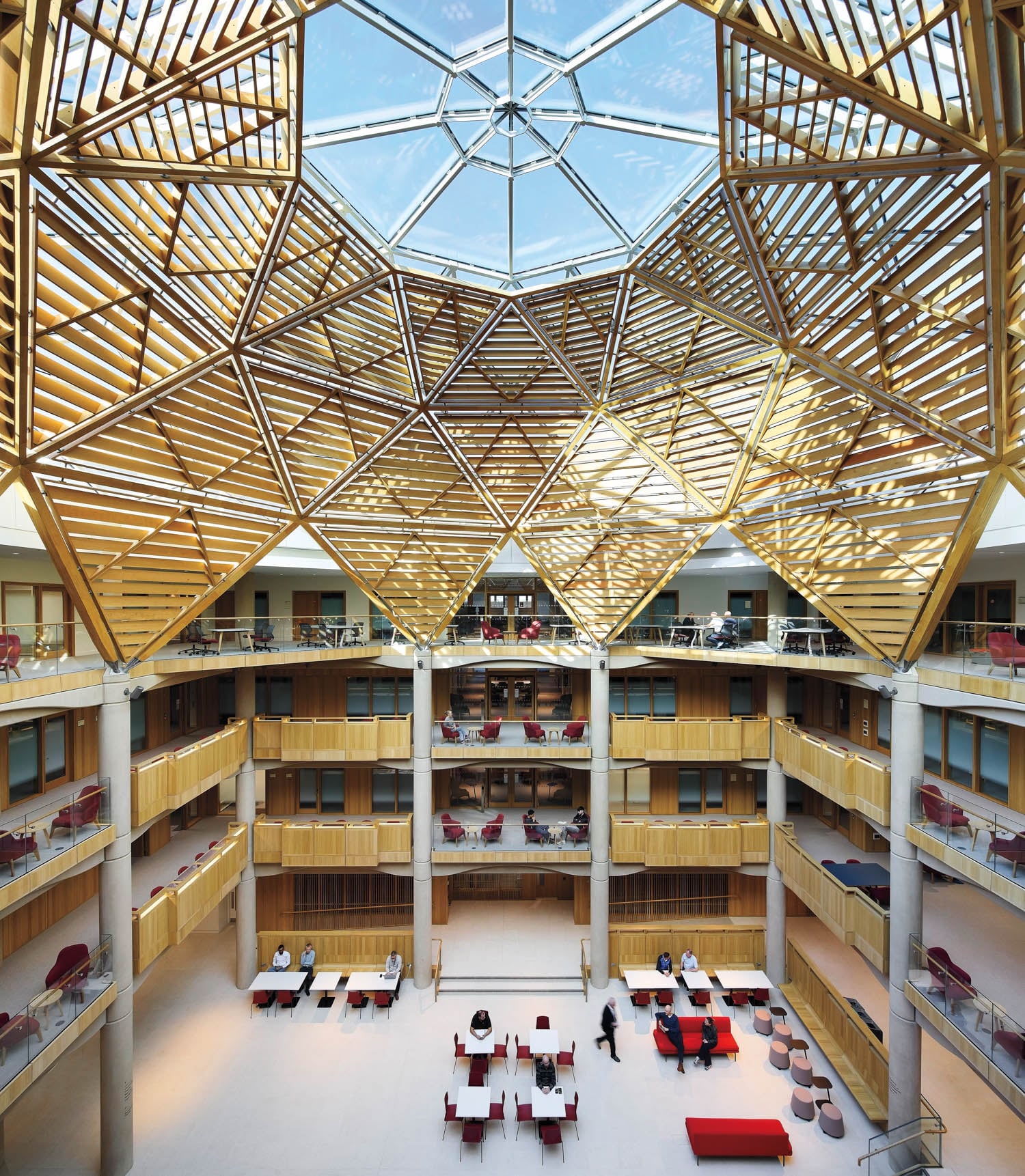 Spacious atrium with multiple balconies, a geometric wooden ceiling structure, and a large glass skylight; tables and chairs are arranged on the ground floor.