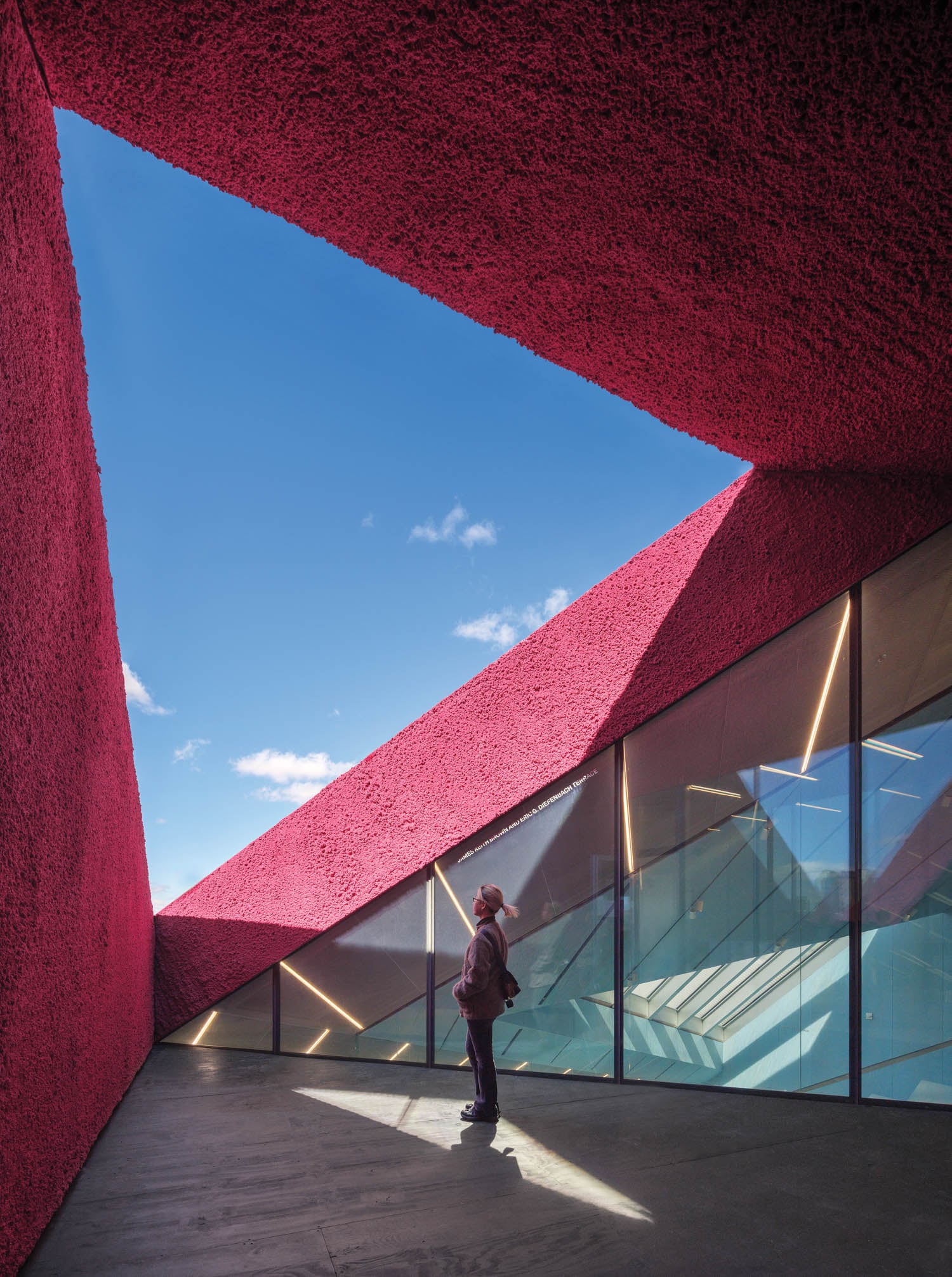 A person stands on a shaded walkway beside glass walls, surrounded by angular red walls opening to a blue sky.