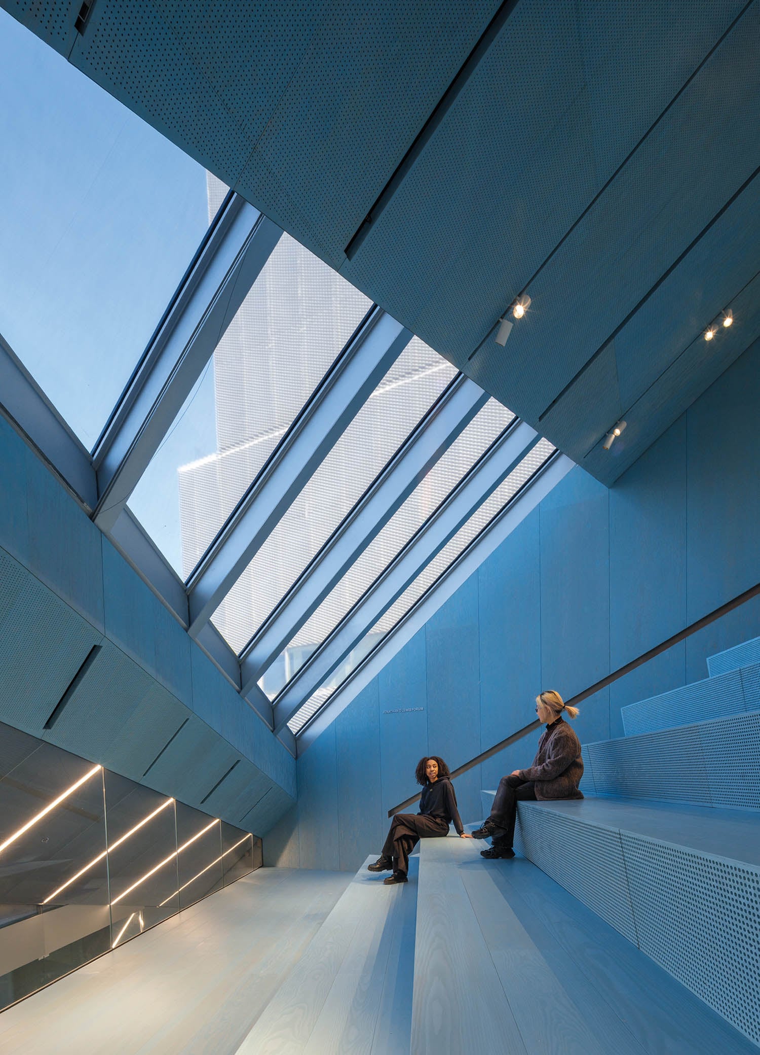 Two people sit on wide, light blue stepped seating beneath large slanted windows in a modern, minimalist interior with views of a city building outside.