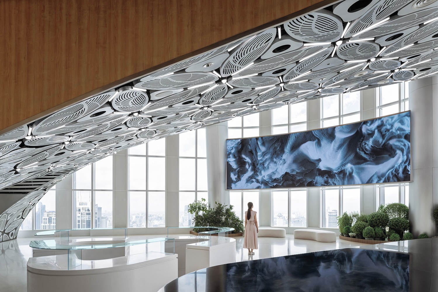A woman stands in a modern, spacious lobby with large windows, an intricate geometric ceiling, and a large digital screen displaying abstract art.