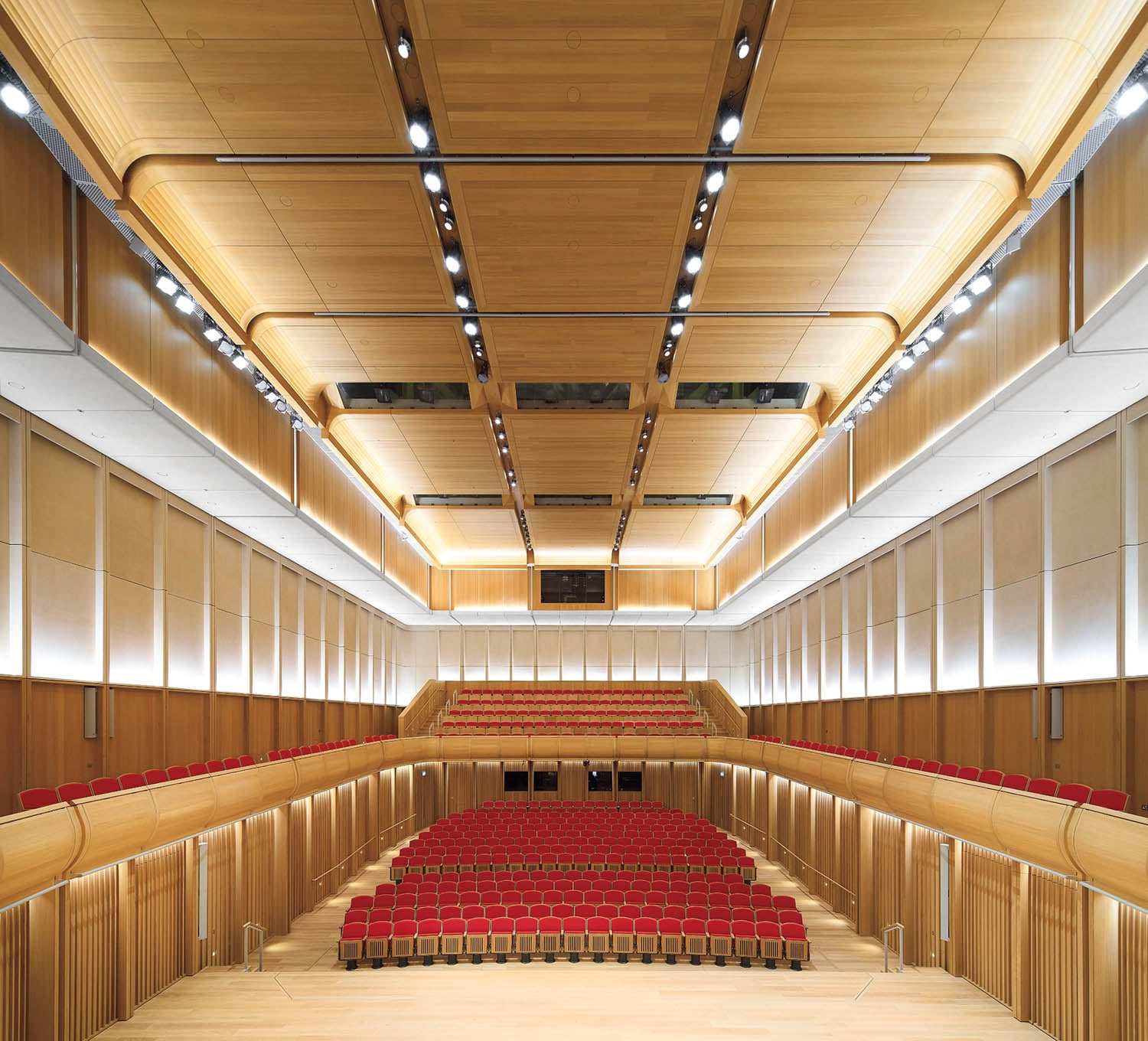 Large modern auditorium with wooden walls and ceiling, red upholstered seats, and balcony seating. The view is from the stage facing the audience.