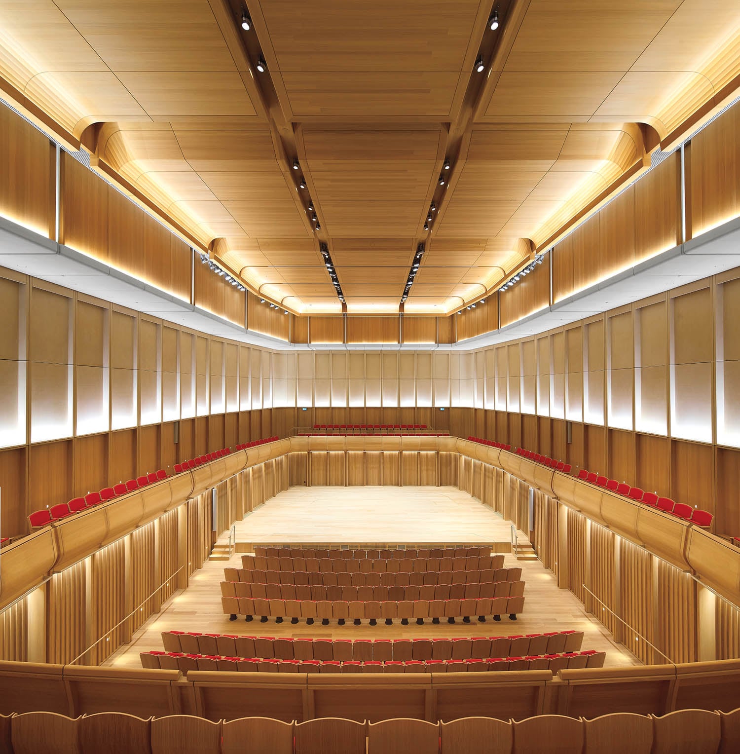 A modern concert hall interior with wooden paneling, multiple rows of red seats, and a large, empty stage under bright lighting.