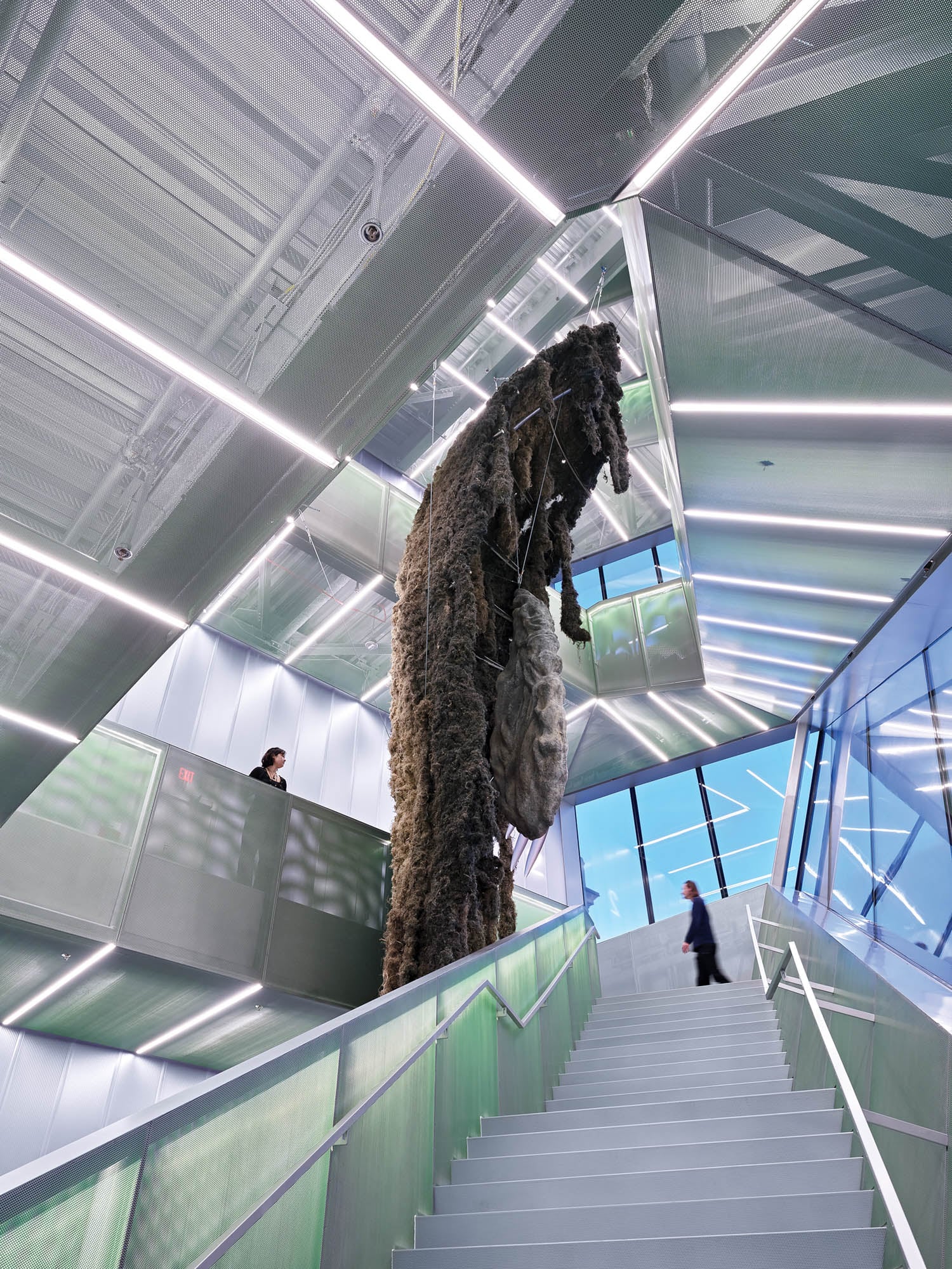 A large, textured sculpture hangs vertically in the center of a modern, glass-walled staircase as two people walk on different levels.