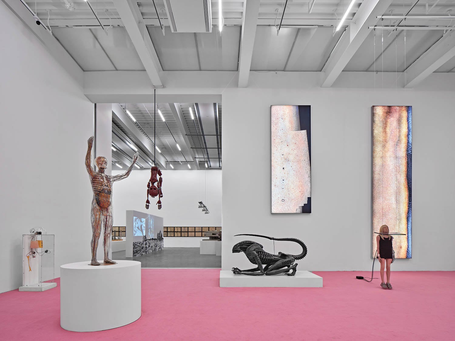 Contemporary art exhibit with anatomical sculpture, suspended figure, abstract wall art, black creature sculpture, and child mannequin on a pink floor in a white gallery space.