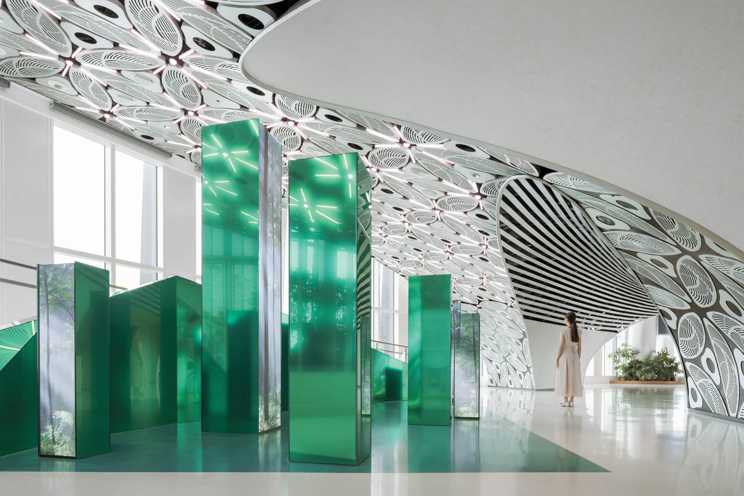 A modern interior with green glass columns, a patterned ceiling, white floors, and a person standing near large windows with natural light.