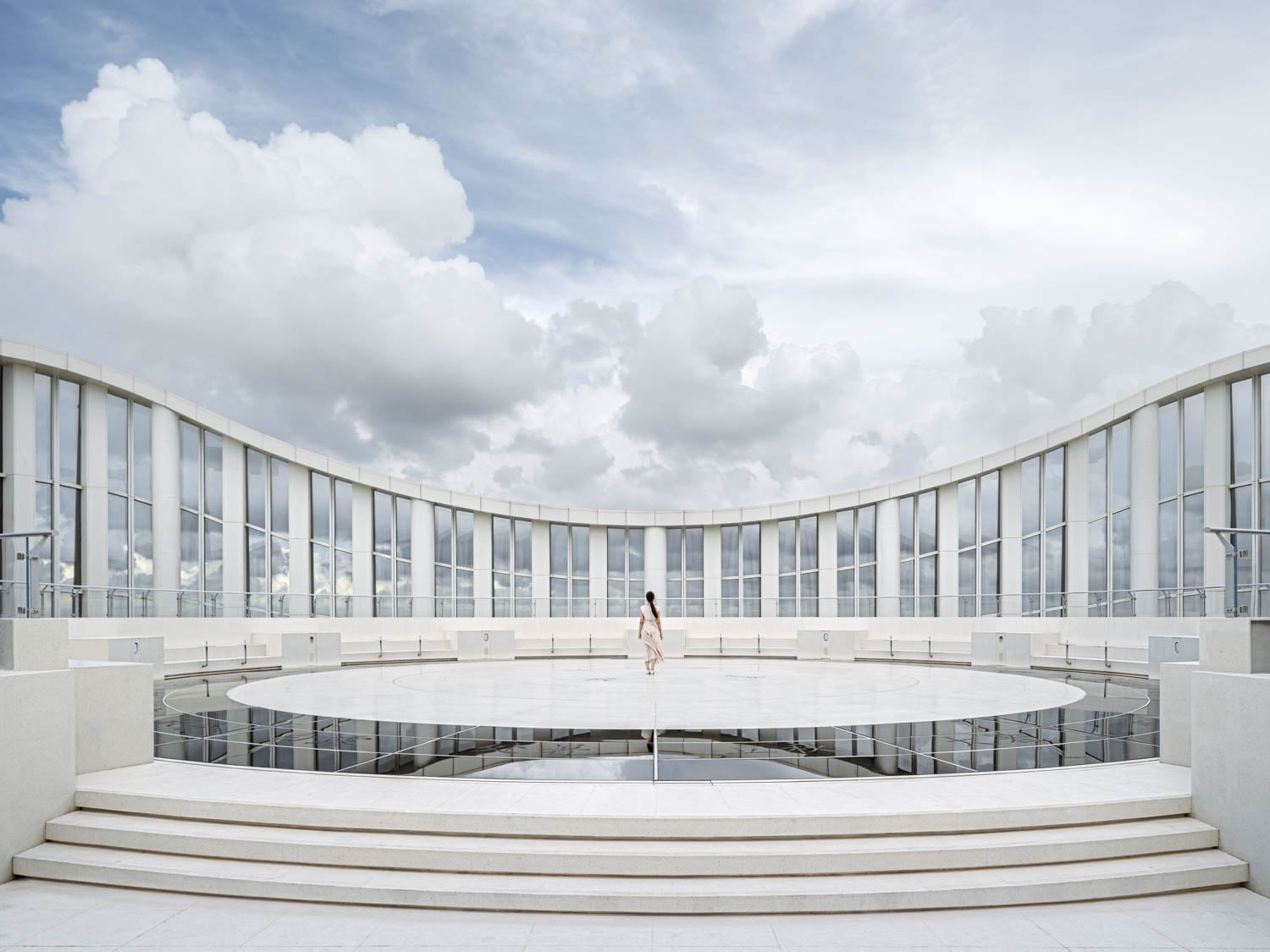 A person stands alone on a large circular platform surrounded by curved modern glass architecture under a cloudy sky.