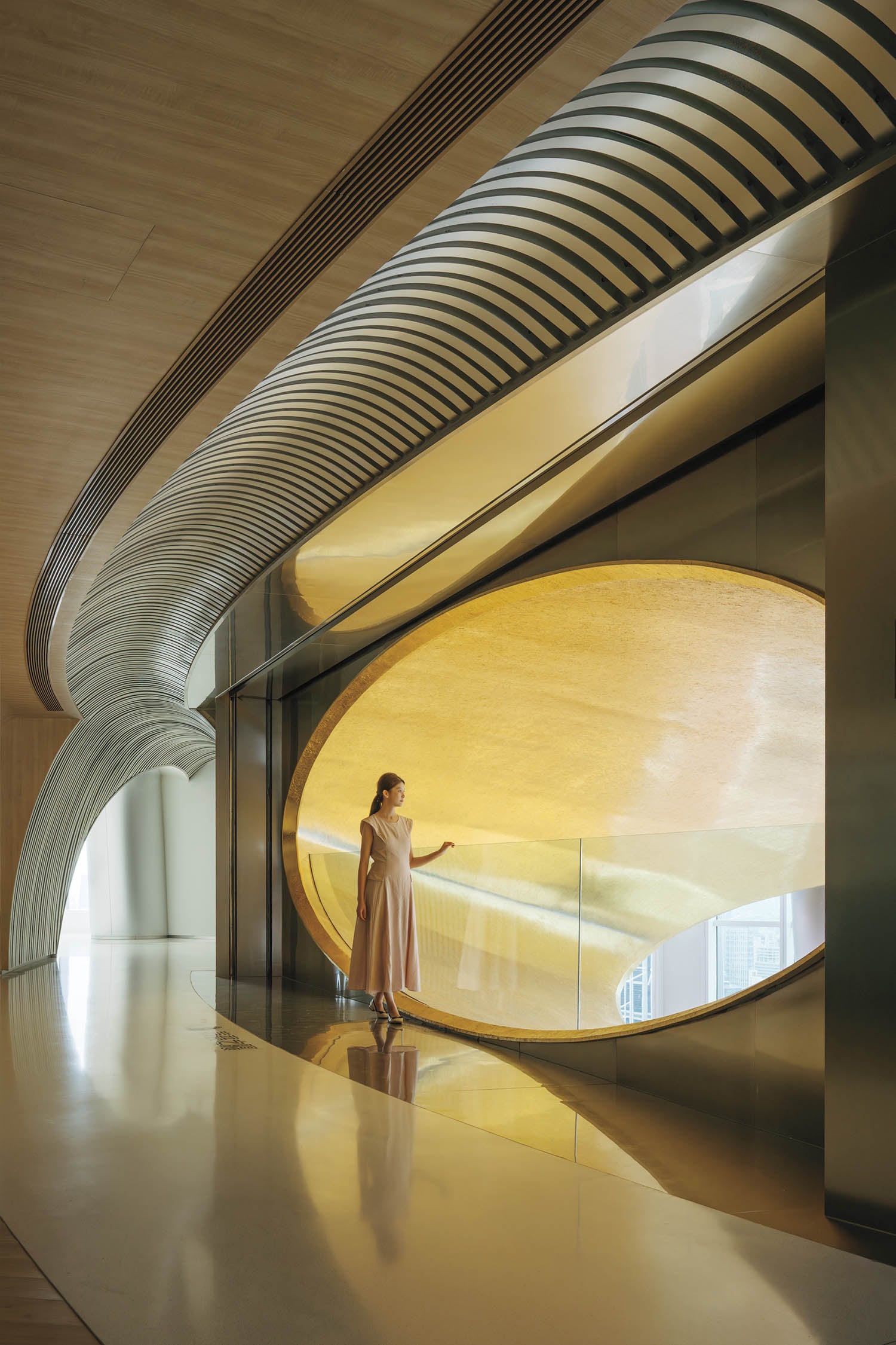 A person in a long beige dress stands in a modern interior with curved architecture and a large yellow oval window feature.