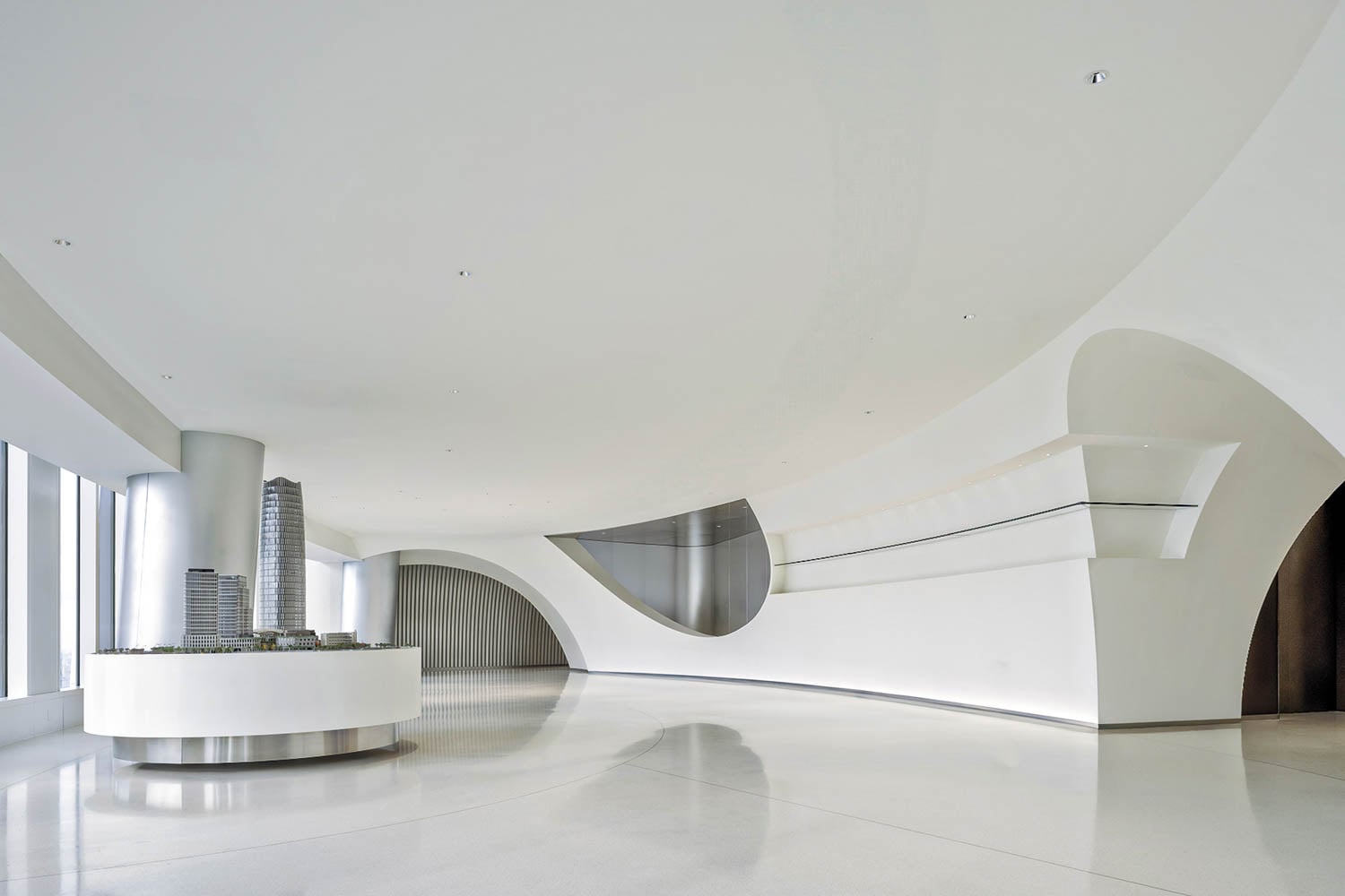 A modern, minimalist interior with curved white walls, large windows, and a circular display featuring a scale model of buildings.