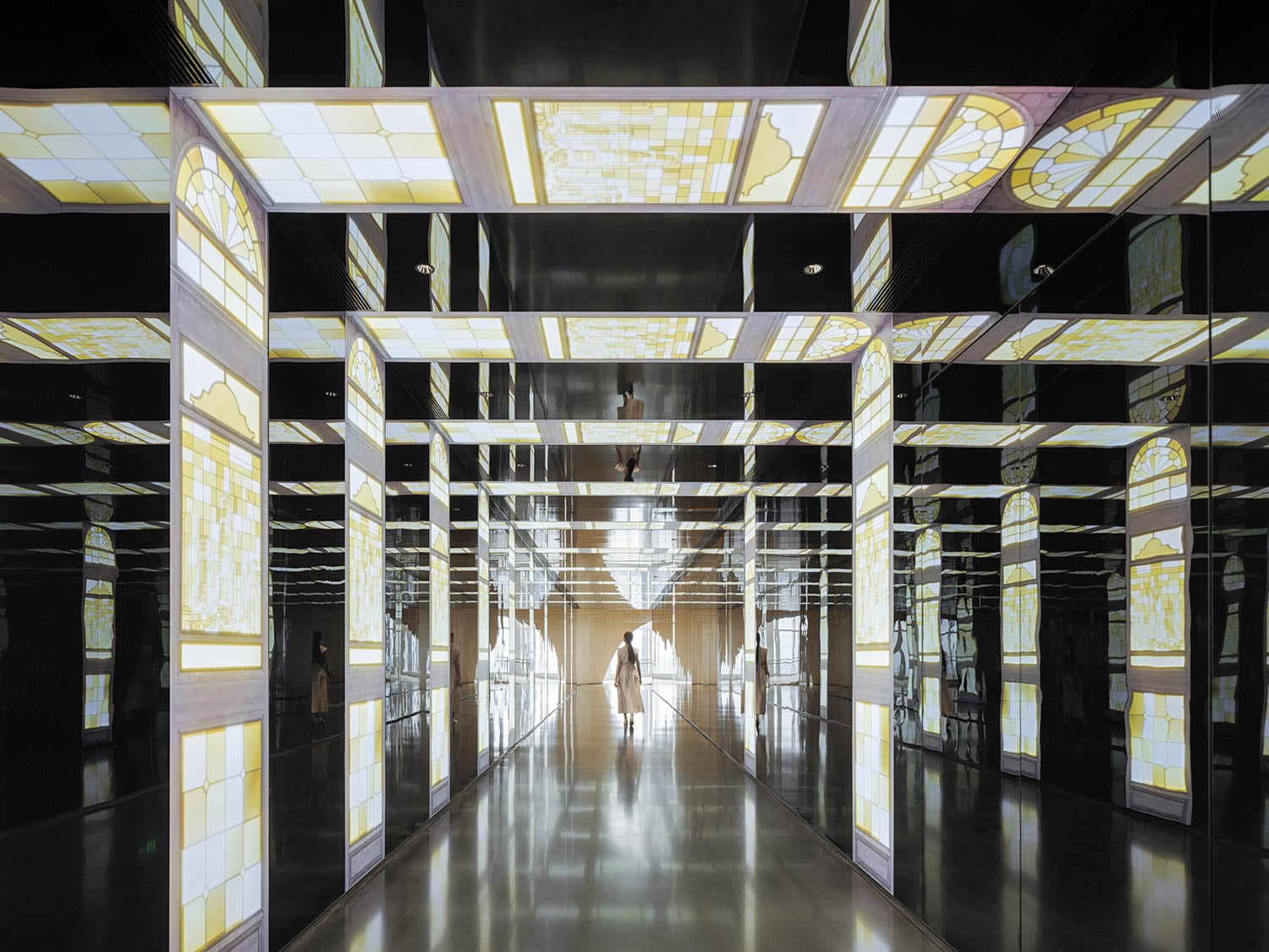 Person walking through a mirrored hallway with illuminated stained glass-like panels on the walls, ceiling, and floor, creating a reflective and geometric environment.