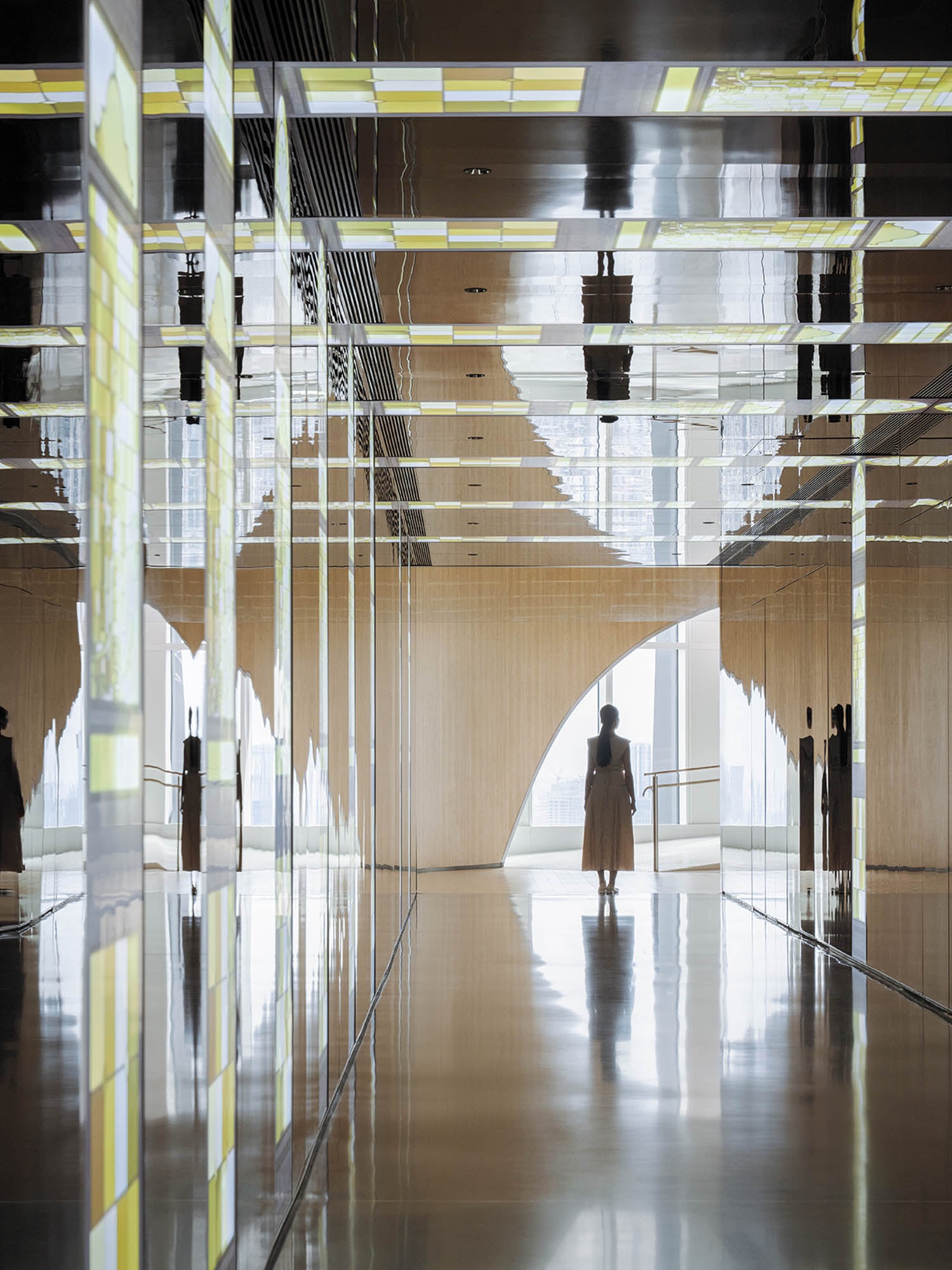 A person stands in a modern, reflective hallway with mirrored walls and a large, curved window in the background letting in natural light.