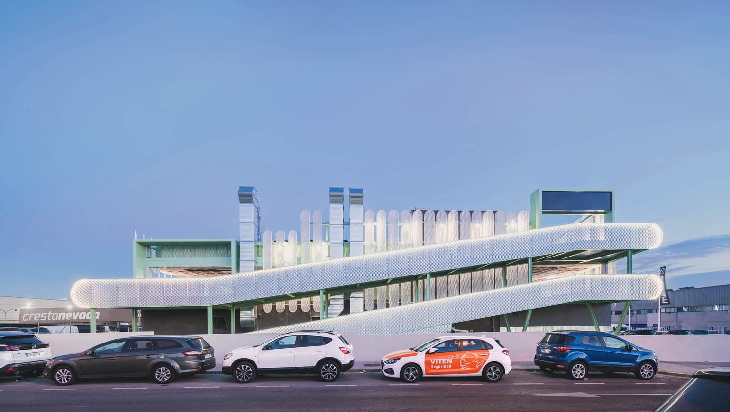 A modern building with a curved, illuminated white ramp and green structural elements, with several parked cars in front on the street.