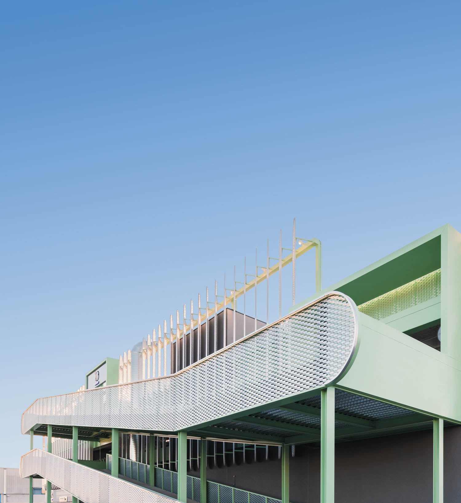 A modern green building with curved metal mesh railings and vertical decorative rods on the roof, set against a clear blue sky.