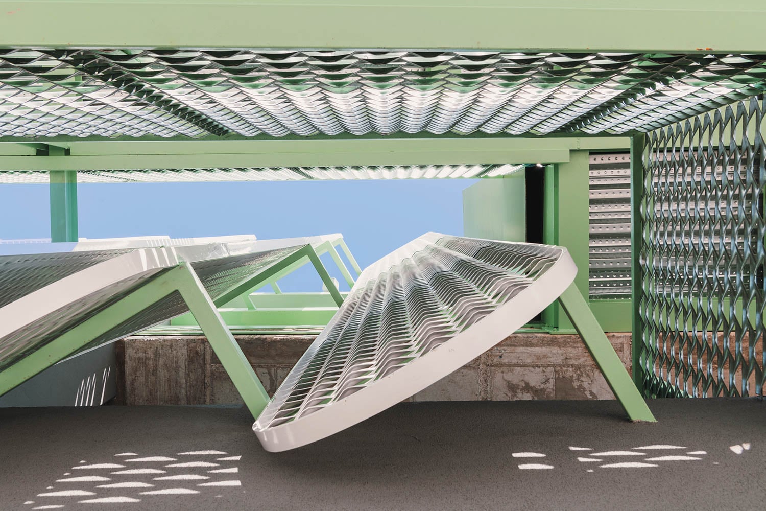 Close-up view of a metal structure with green frames and perforated panels, some of which are tilted open, against a clear blue sky.