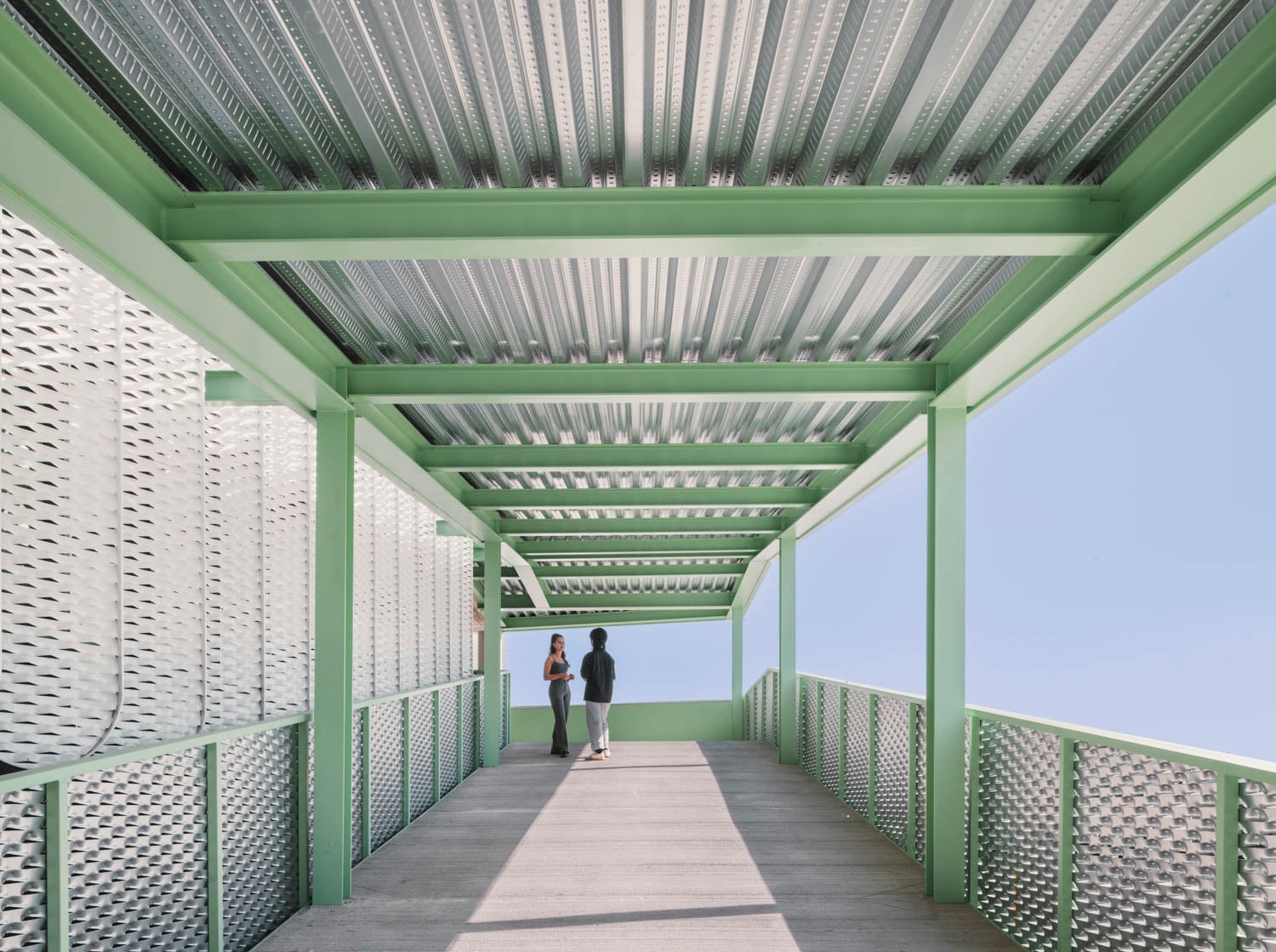 Two people stand talking on a covered outdoor walkway with green metal beams and perforated metal walls, under a clear blue sky.