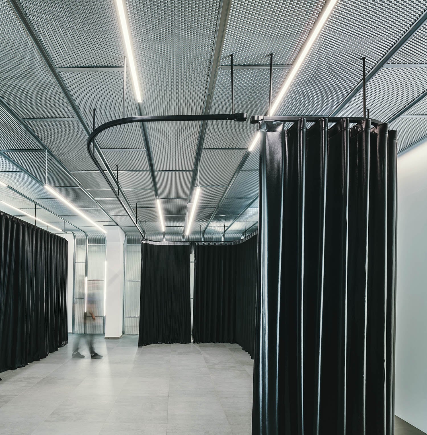 A modern interior space with metal grid ceiling, bright linear lighting, gray tiled floor, and black privacy curtains; a blurred person walks in the background.