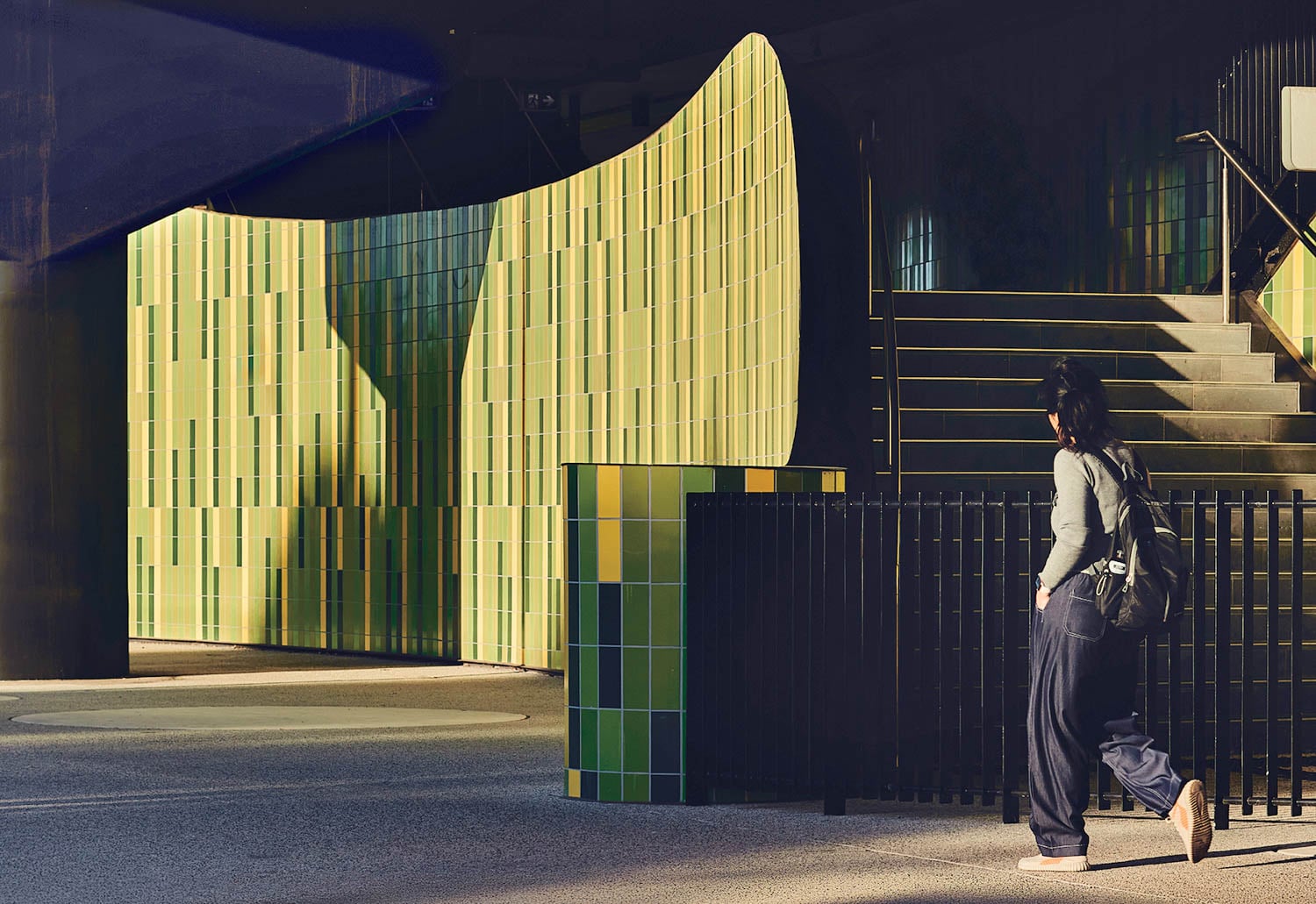 Person walking past a curved wall of yellow-green tiles and a staircase in an urban setting, with strong sunlight casting shadows.