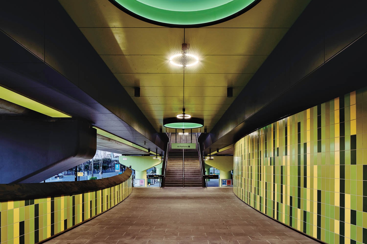 Modern subway station interior with green and yellow tiled walls, central staircase, overhead circular lights, and a curved corridor leading to the platform.