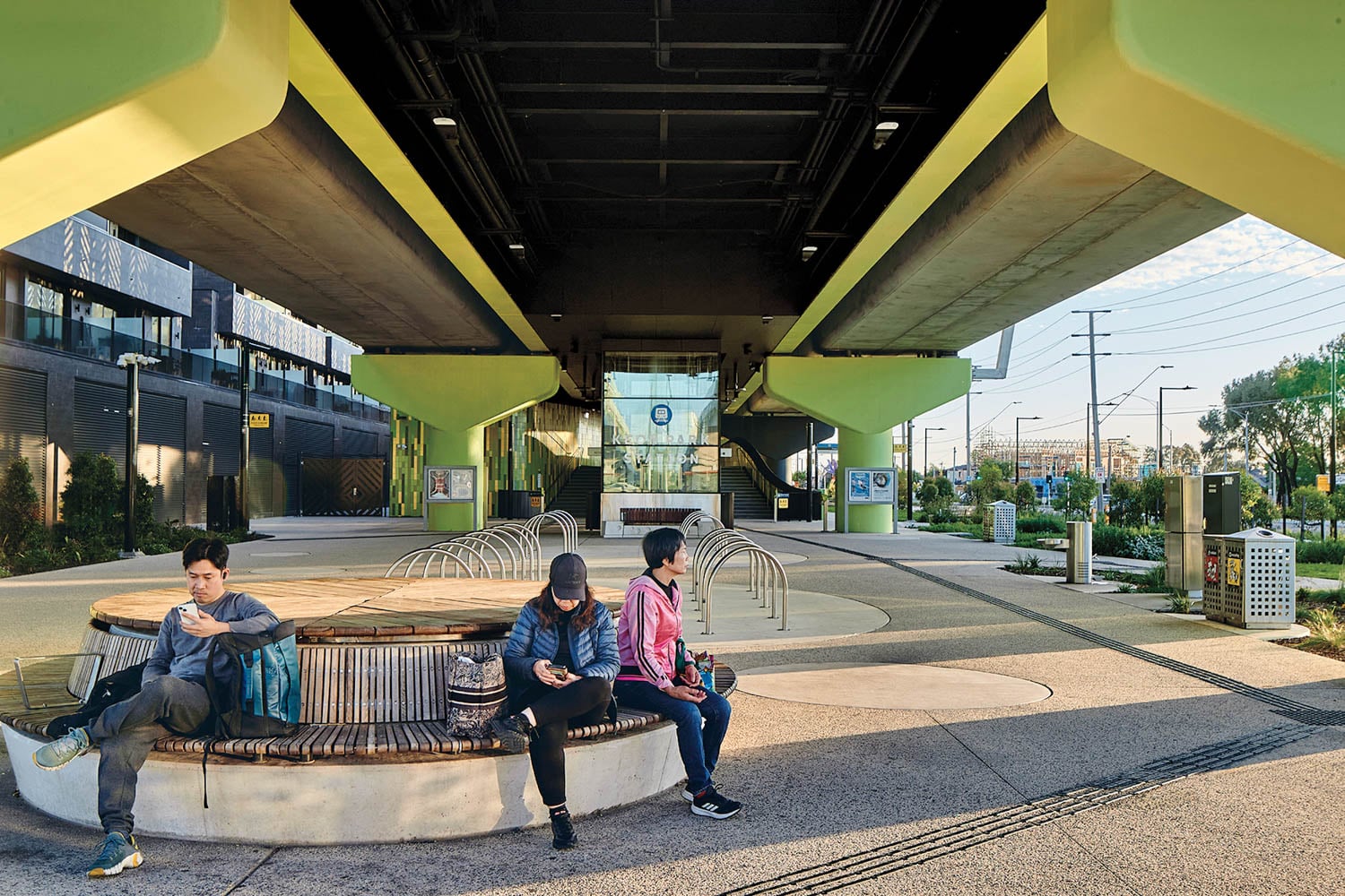 Three people sit on a curved bench under an elevated train track in an urban public space, looking at their phones.