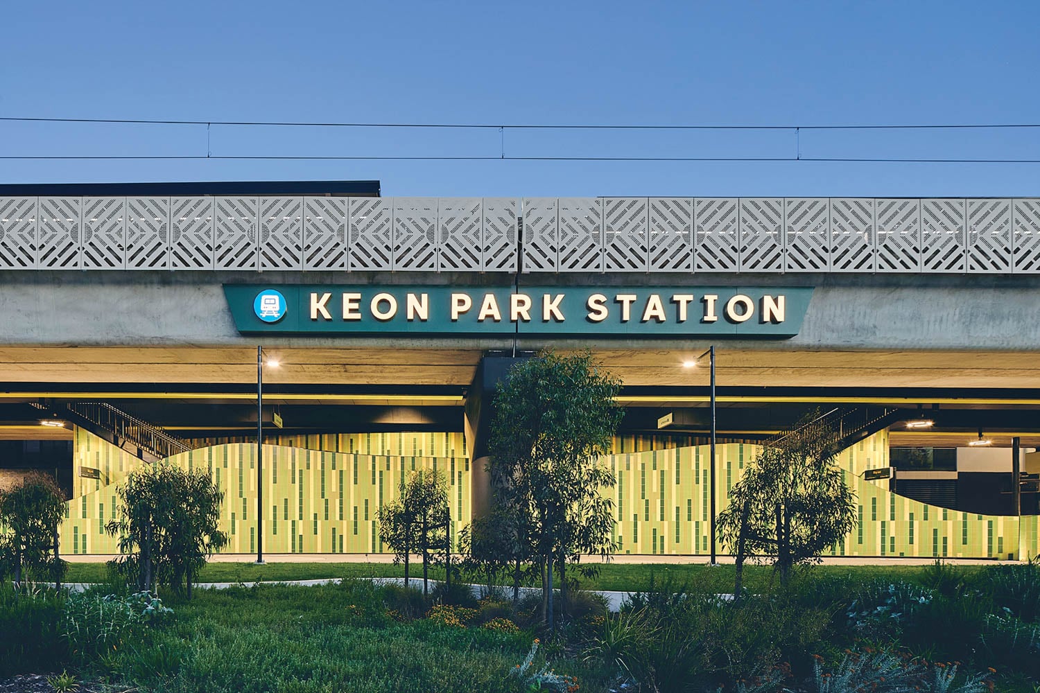 Exterior view of Keon Park Station, showing its illuminated sign, modern facade with patterned panels, and landscaped surroundings at dusk.