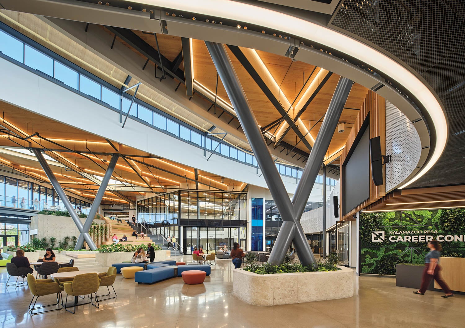 Modern, spacious atrium with large windows, exposed beams, seating areas, indoor plants, and a sign reading “Kalamazoo Area Career Ctr.” Several people are sitting and conversing.