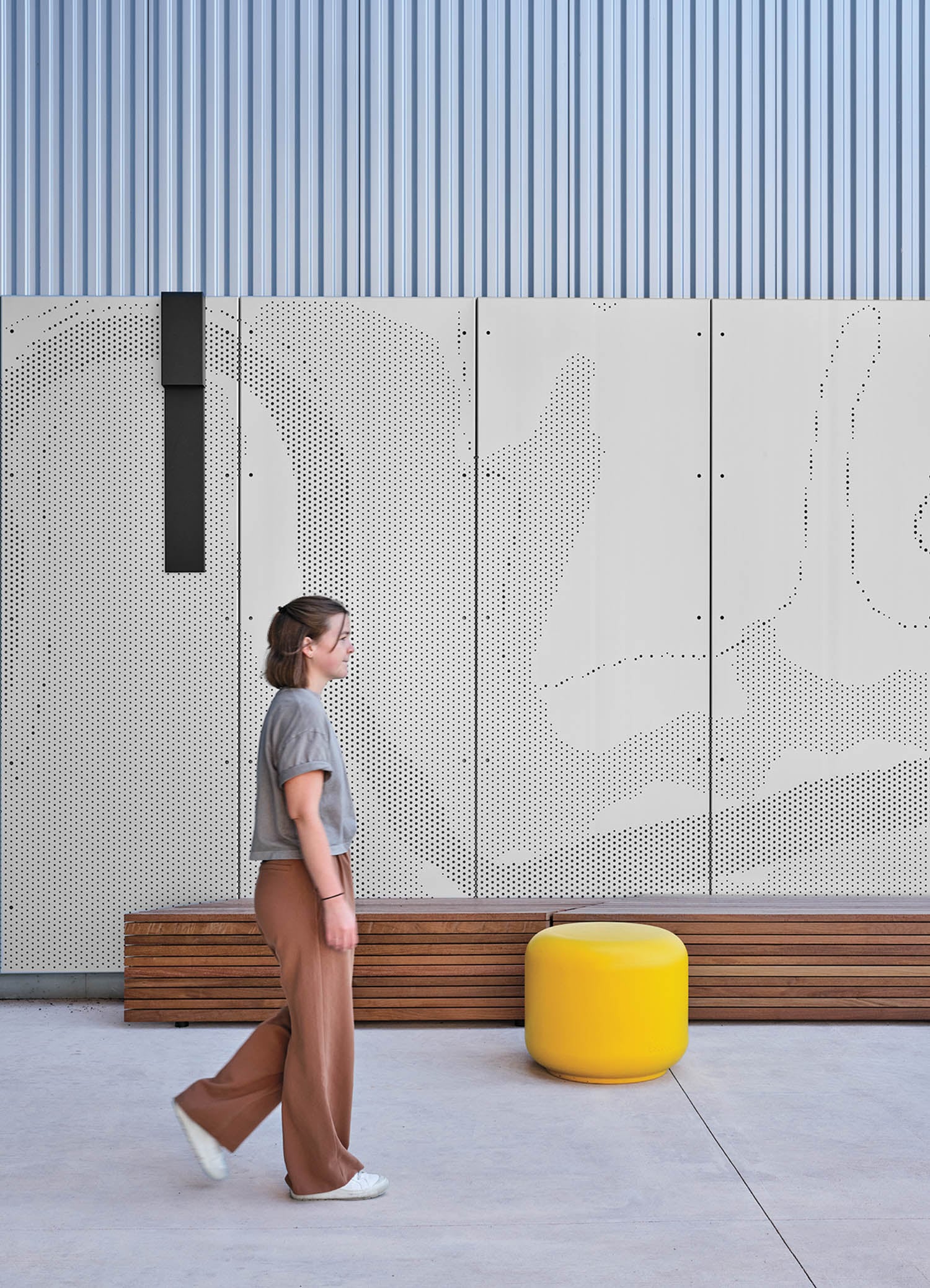 A person walks past a perforated metal wall with abstract patterns, a wooden bench, and a yellow round ottoman in a modern outdoor setting.