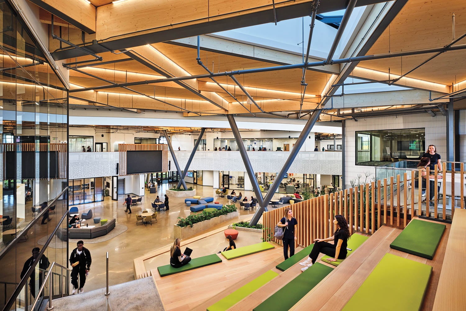 Modern office interior with open seating areas, glass walls, wooden accents, and people working or walking throughout the spacious, brightly lit atrium.