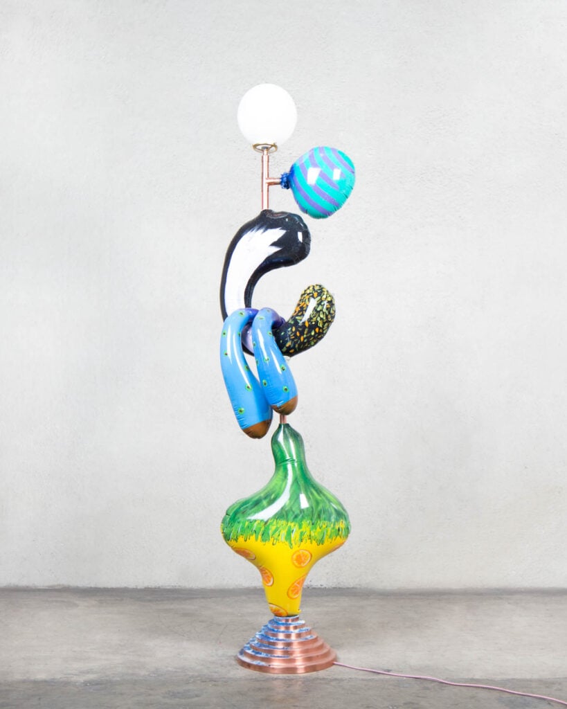 A colorful, abstract floor lamp with a round white bulb, curved blue and black sections, and a green and yellow base, standing against a plain gray wall.