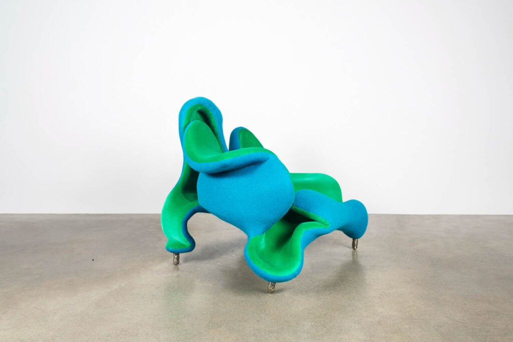 A sculptural chair with bold, curving forms in bright blue and green upholstery sits on a concrete floor against a plain white wall.