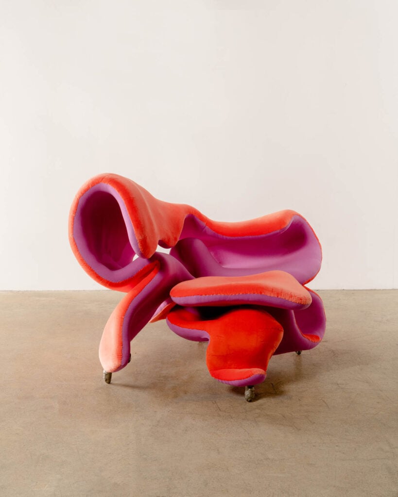 A sculptural, abstract chair with curved, organic shapes in red, pink, and purple fabric, set on a plain concrete floor against a white wall.