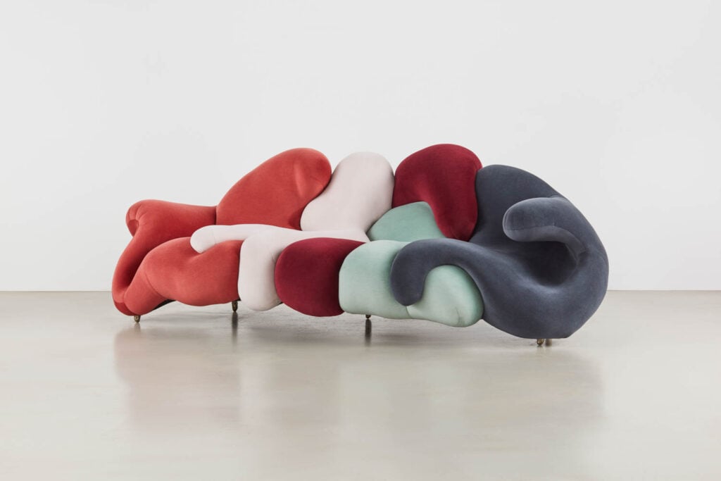 A multicolored, abstract-shaped sofa with curved, padded sections in red, pink, green, and dark blue sits on a light floor against a plain white wall.