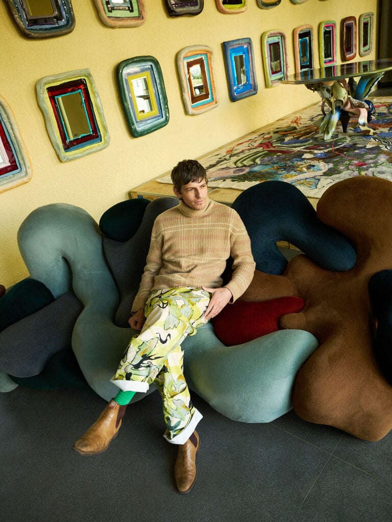 A man wearing a beige sweater, patterned pants, and brown boots sits on a sculptural, multicolored sofa in a room with colorful framed mirrors on the wall.