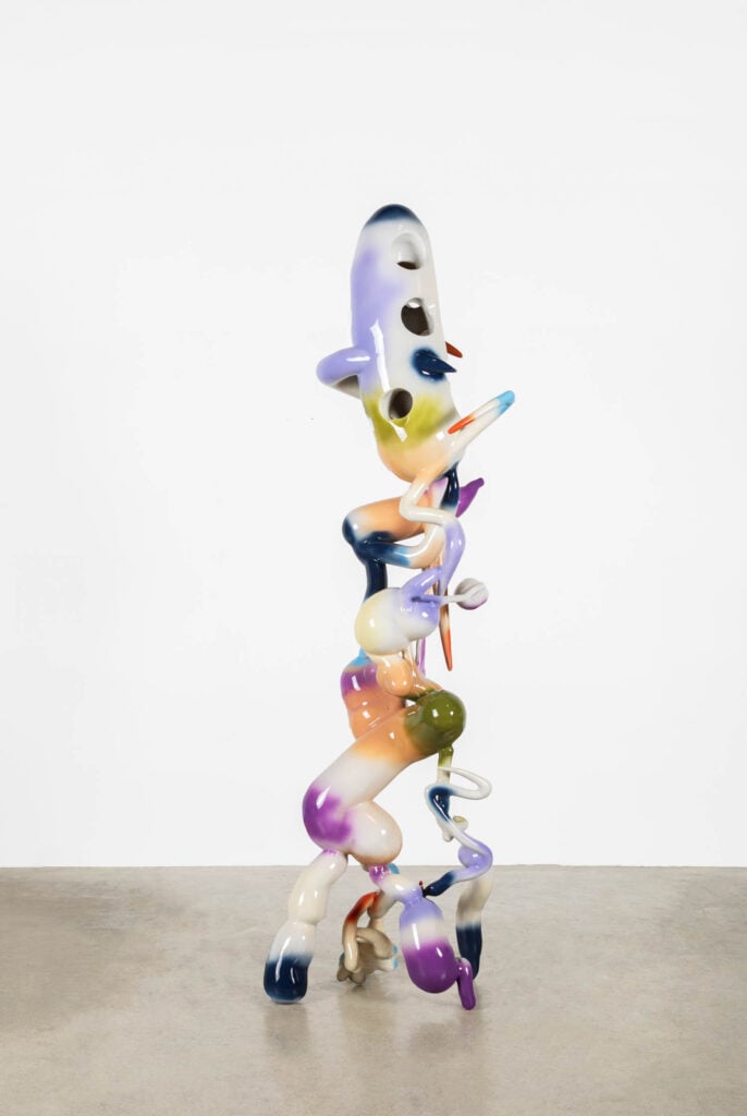 Tall, abstract sculpture with a glossy, multicolored surface and irregular, bulbous shapes, standing on a plain concrete floor against a white background.