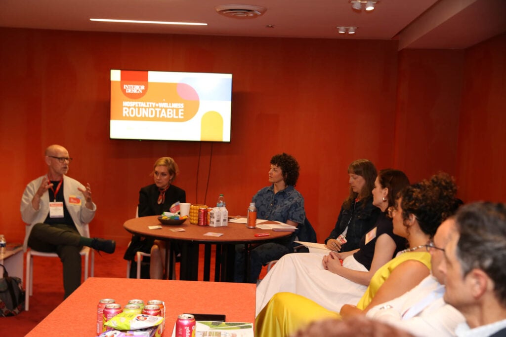 A group of people sit around a table in discussion at a “Hospitality + Wellness Roundtable” event, with snacks and drinks on the table and a presentation screen in the background.