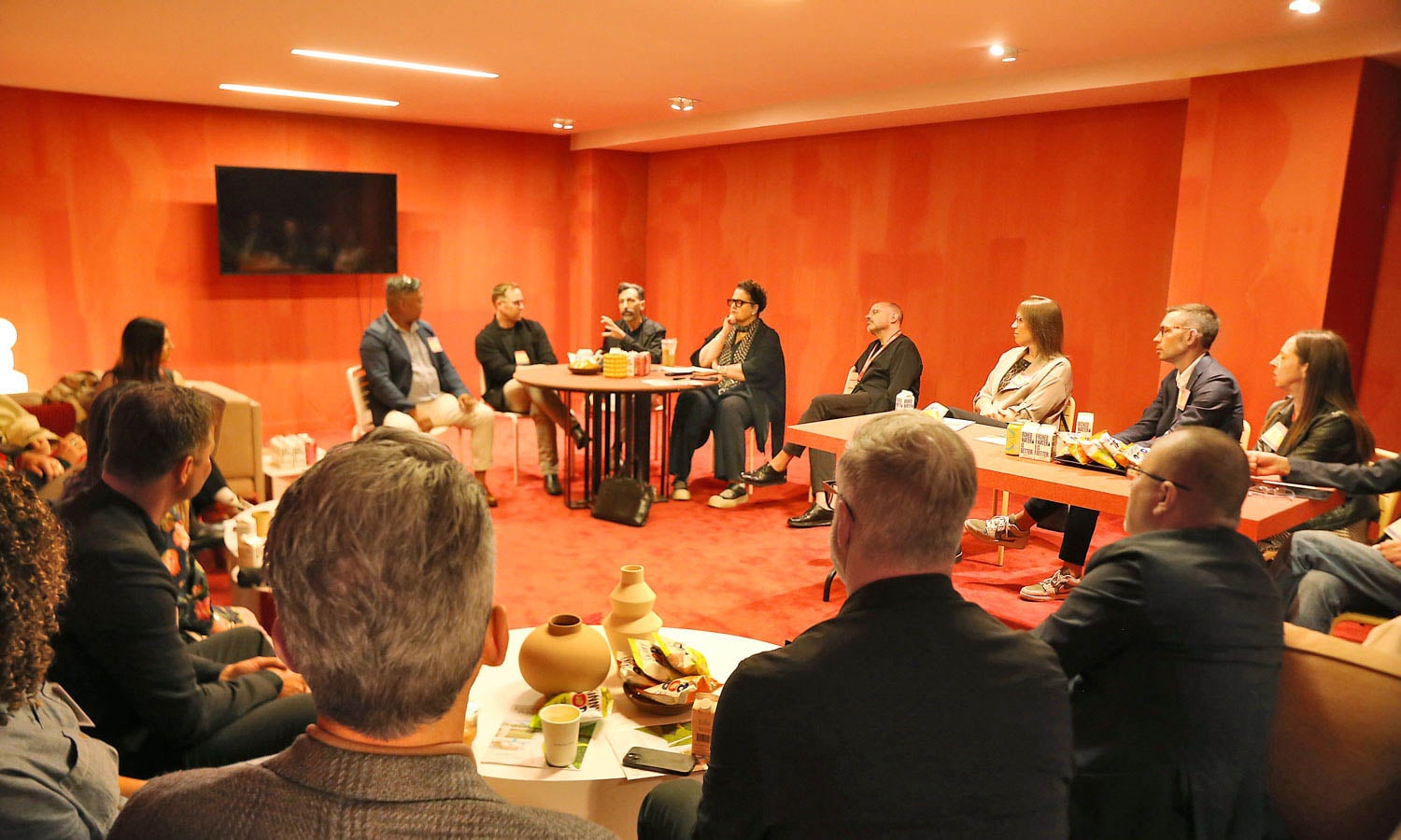 A group of people sit in a red-walled meeting room, arranged in a circle, listening to a speaker; snacks and drinks are on tables.