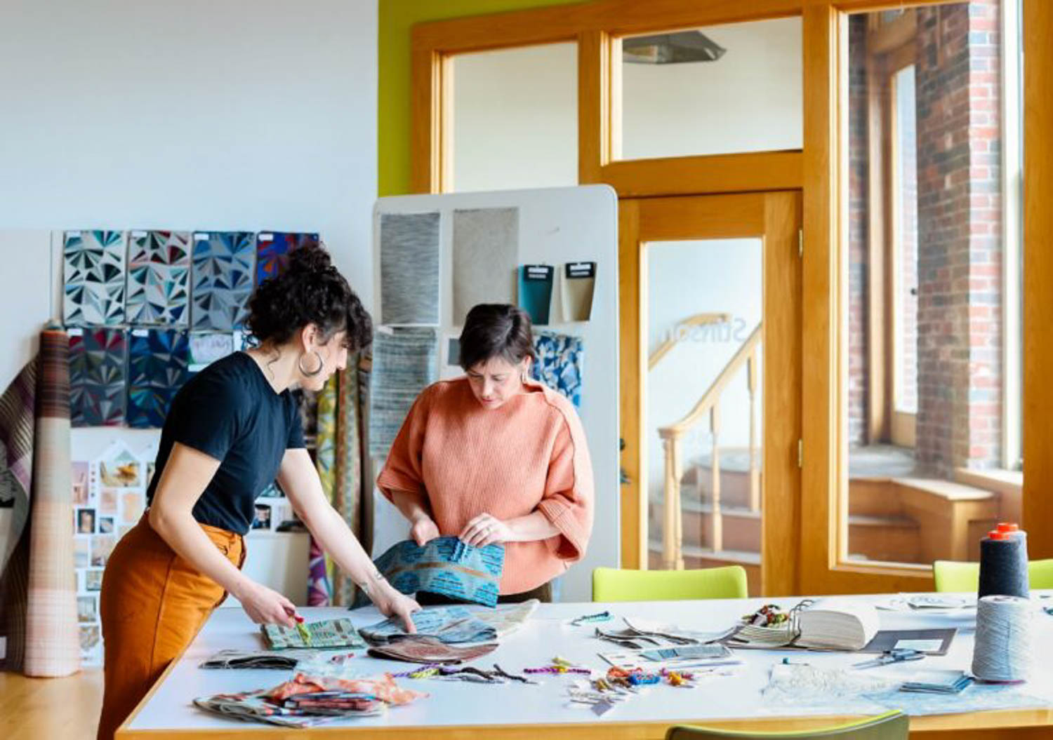 Two people stand at a table covered with fabric swatches and samples, examining materials in a brightly lit room with textile displays on the wall.