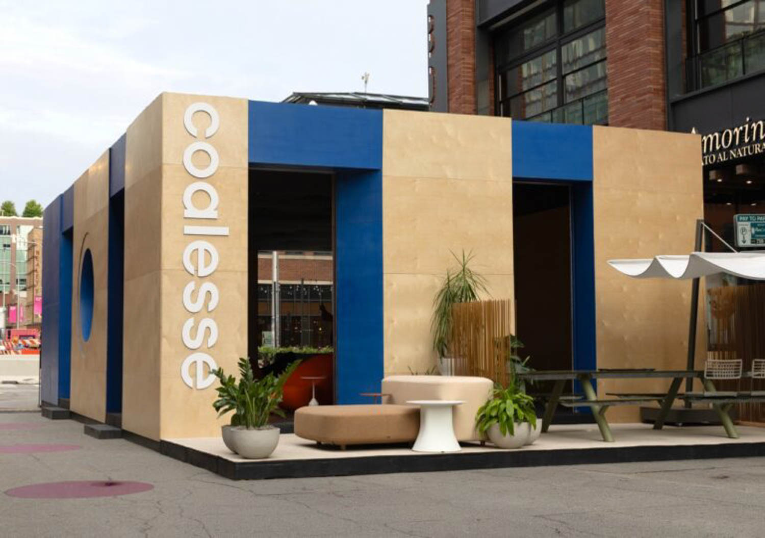 A modern, box-shaped building with tan and blue exterior panels displays the word "Coalesse" vertically; outdoor seating and plants are arranged in front.