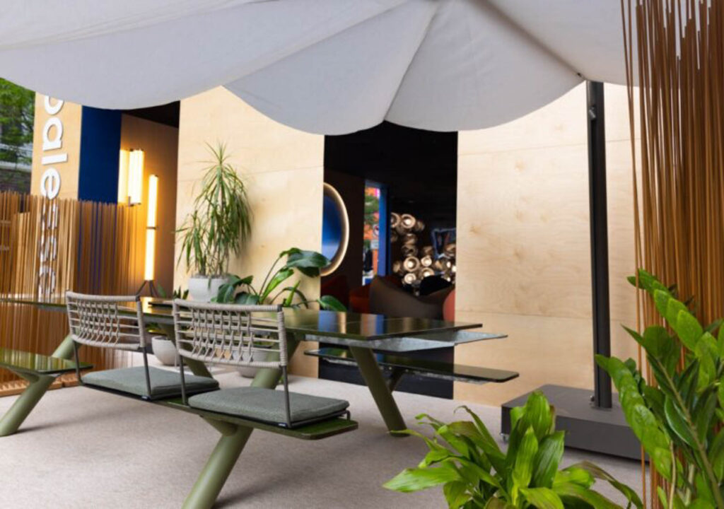 Modern outdoor seating area with a green metal table, wire-frame benches, potted plants, and a large white umbrella, set against a wooden wall backdrop.