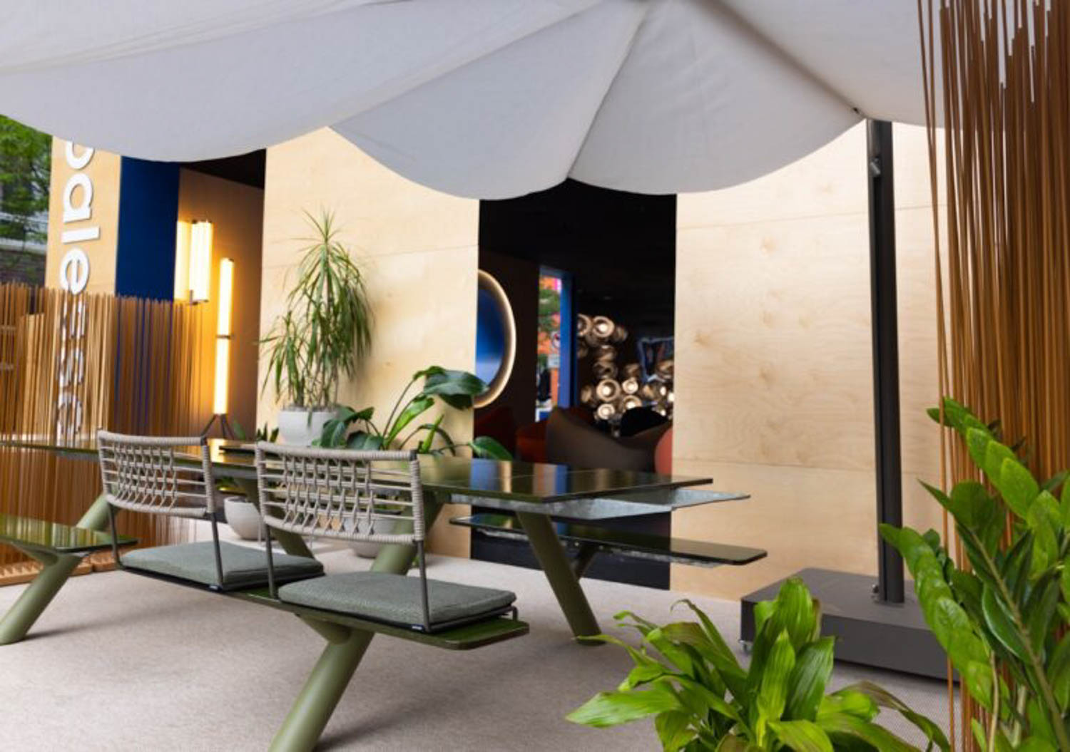 Modern outdoor seating area with a green metal table, wire-frame benches, potted plants, and a large white umbrella, set against a wooden wall backdrop.
