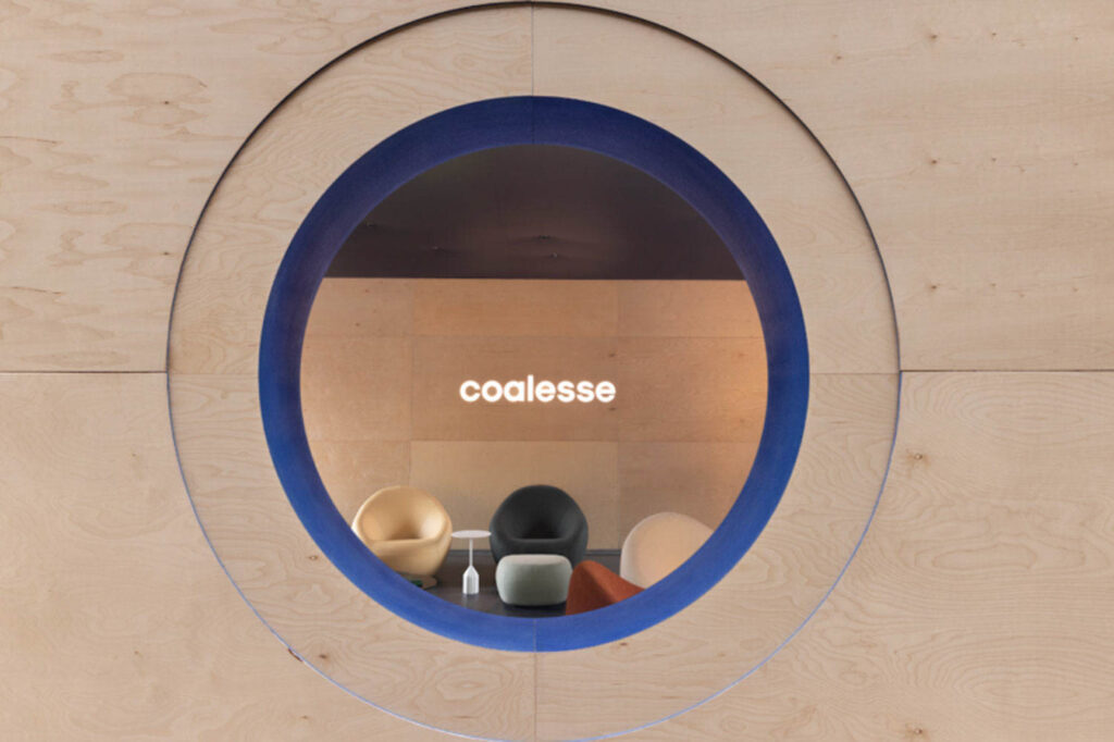 A round window in a wooden wall frames a minimalist lounge with modern chairs and a glowing "coalesse" sign on the back wall.