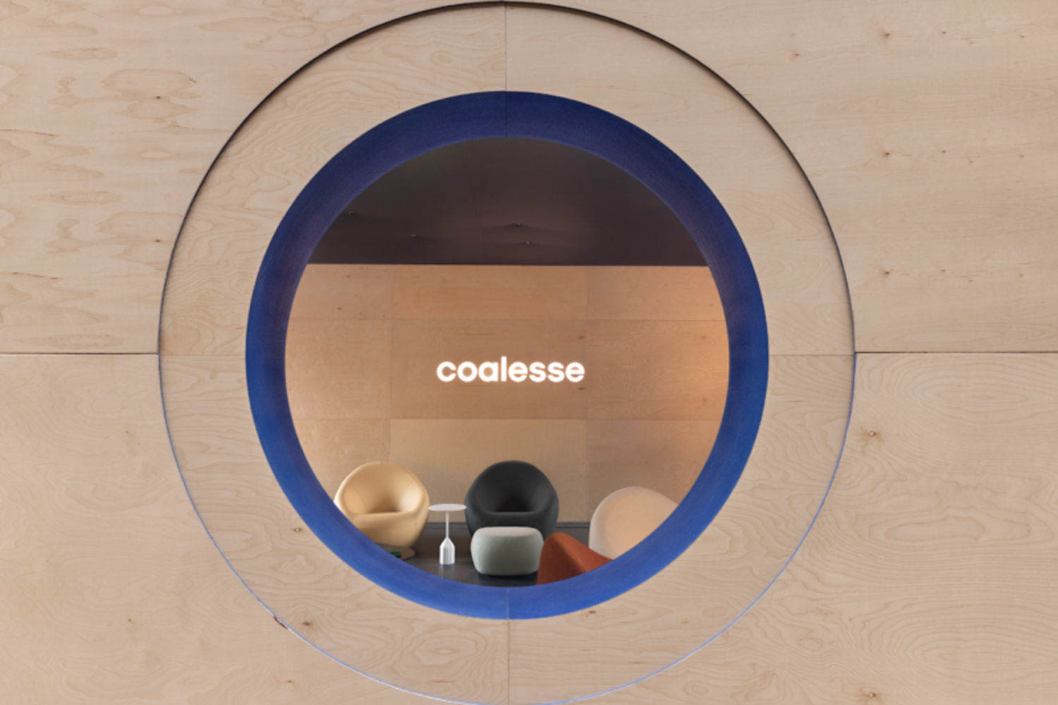 A round window in a wooden wall frames a minimalist lounge with modern chairs and a glowing "coalesse" sign on the back wall.