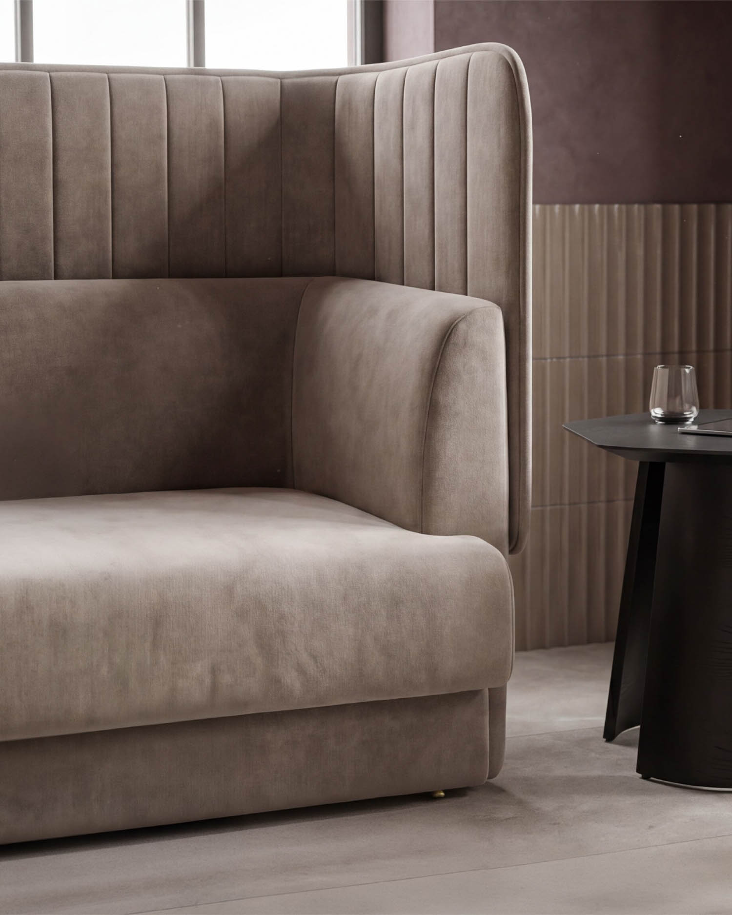 A close-up of a modern beige upholstered sofa with vertical stitching near a black side table holding a glass of water, set against tiled and painted walls by a window.