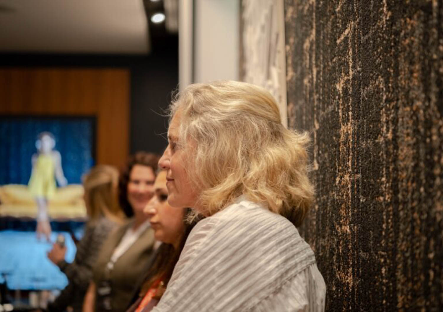 Several women stand indoors near a textured wall, with one woman in the foreground facing to the right. A blurred television screen and other people are visible in the background.