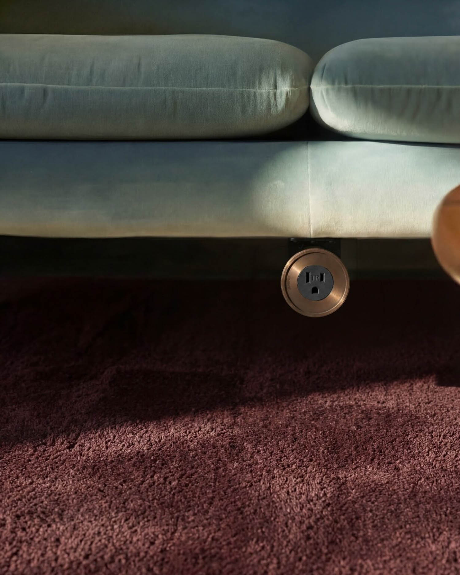 A close-up of a mauve carpet and the bottom of a light green couch with a round electrical outlet installed on the couch’s lower frame.