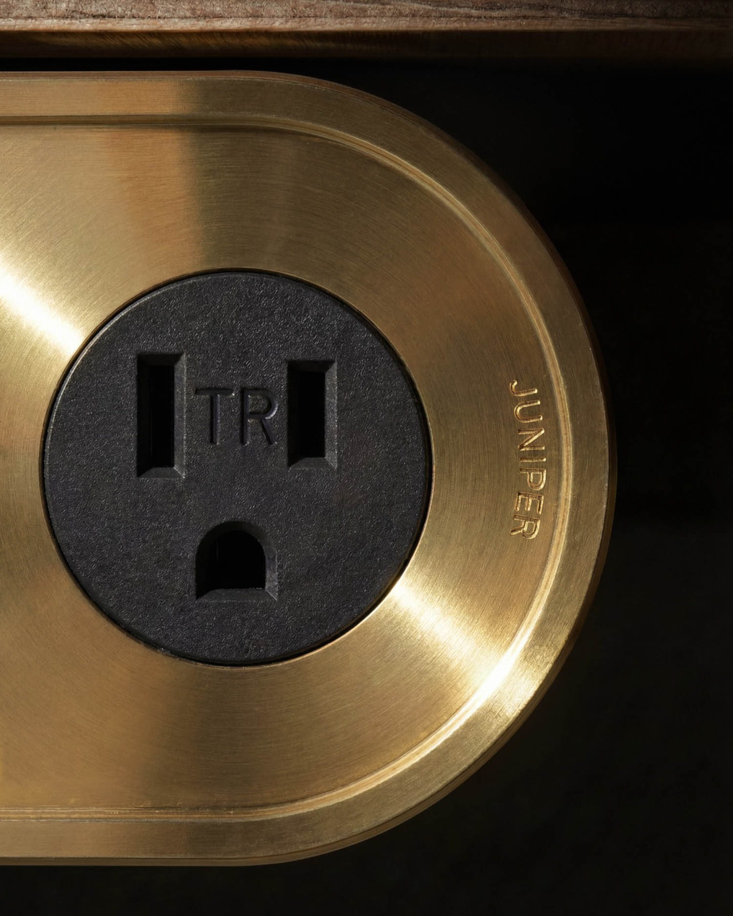 Close-up of a brass electrical outlet plate with a black outlet labeled "TR" and the word "JUNIPER" engraved on the metal surface.