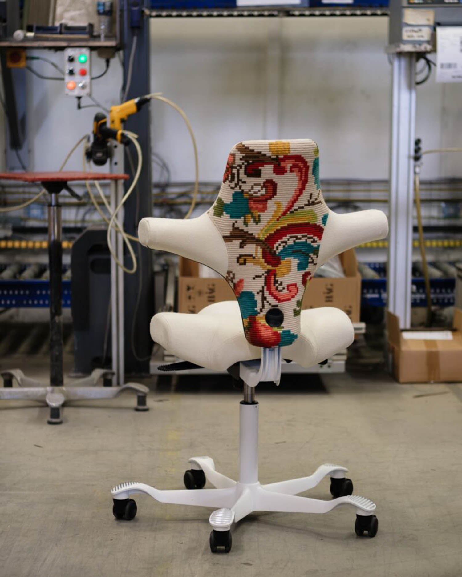 An office chair with a white base and armrests, featuring a colorful, patterned backrest, is positioned in an industrial workspace with tools and machinery in the background.